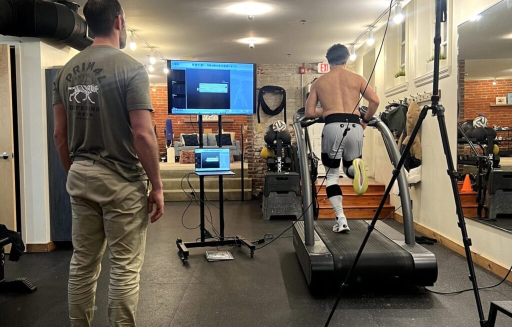 "Shirtless man running on a treadmill while a therapist performs gait analysis, observing his form and monitoring his movement closely.